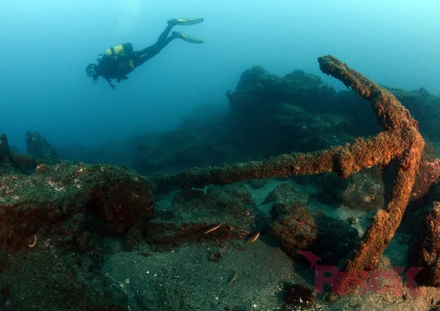 A rusty anchor on the ocean floor with a scuba diver in the background