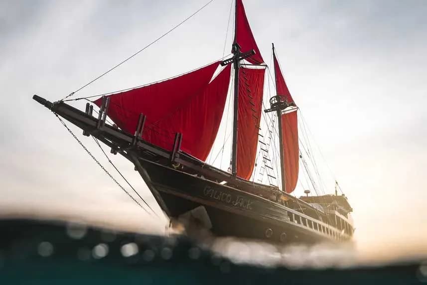 A red sailboat named Calico Jack glides across the ocean with a vibrant sunset illuminating the sky behind it.