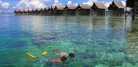 Two people snorkel a coral reef with a resort on stilts in the background. Two people snorkel a coral reef with a resort on stilts in the background.