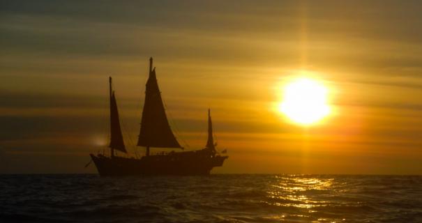 A boat sails calms seas with the sun setting in the background.