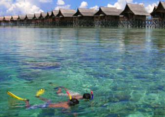 Two people snorkel a coral reef with a resort on stilts in the background.