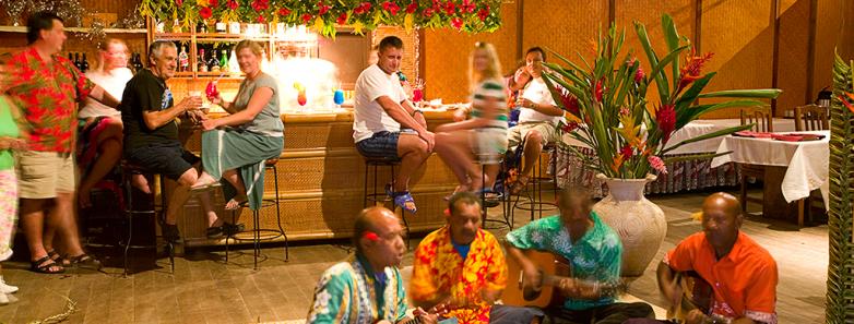 People enjoy drinks at a bar while musicians perform sitting on the ground.