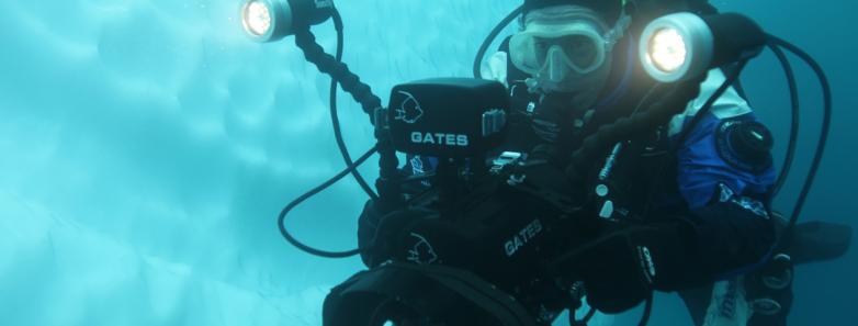 A person holding a large underwater camera system scuba dives in Antarctica