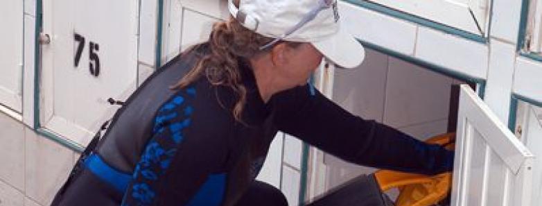 A woman in a wetsuit and hat places personal items in a lockable locker.