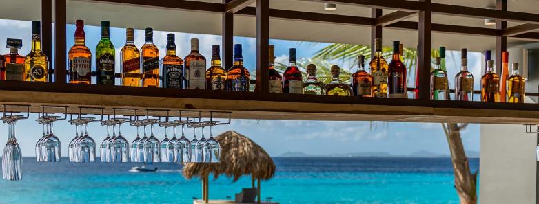 A shelf of bottles of liquor with the sea in the background at Delfins Beach Resort Bonaire.