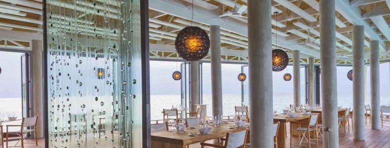 A modern restaurant with a long table and glass walls, offering a spacious and bright dining experience atKuramathi Island Resort Maldives