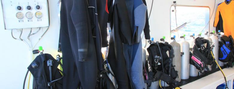 Wetsuits hang in front of full scuba equipment sets on the dive deck of the MV Valentina liveaboard.
