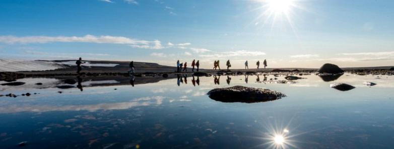 A group of people walk along the edge of a still body of water with the sun in the background